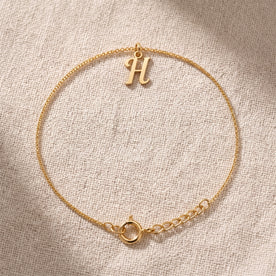 Personalised gold initial bracelet with the letter H in the Bella style, laid flat on linen, showing the whole bracelet.