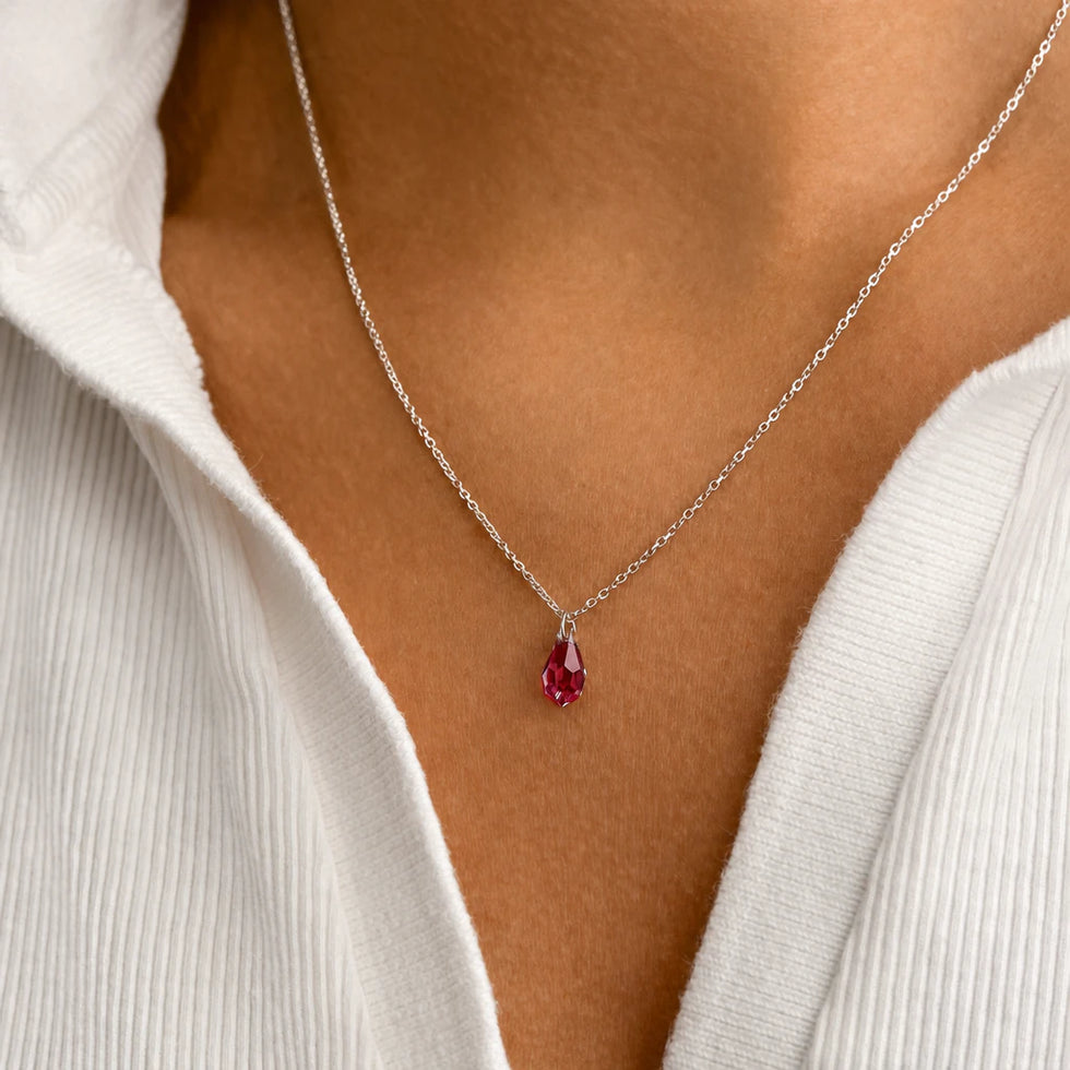 Model wearing a silver birthstone crystal drop necklace with a ruby red teardrop pendant over a white ribbed top